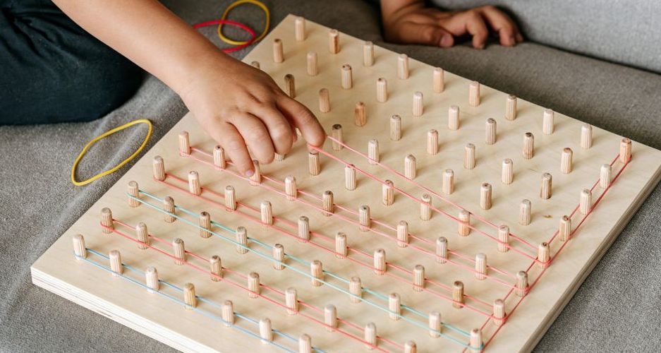Uma criança utiliza elásticos num tabuleiro de pinos, uma estratégia eficaz para fortalecer as mãos e trabalhar a motricidade fina, promovendo controlo, precisão e coordenação dos movimentos.
