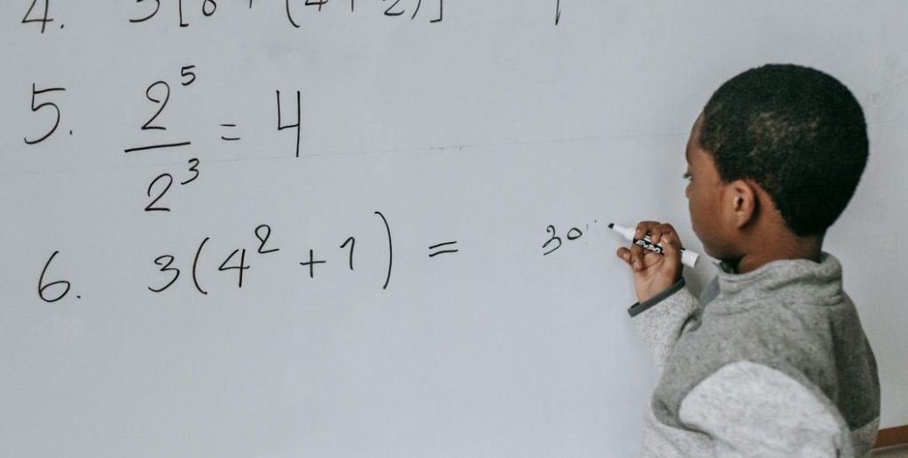 Uma criança resolve operações no quadro com alguma hesitação, evidenciando dificuldades associadas à discalculia, que afetam a compreensão dos números, dos cálculos e das relações matemáticas.