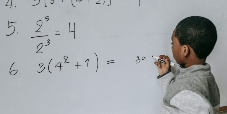 Uma criança resolve operações no quadro com alguma hesitação, evidenciando dificuldades associadas à discalculia, que afetam a compreensão dos números, dos cálculos e das relações matemáticas.