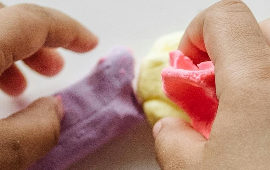 As mãos de uma criança manipulam massa moldável colorida, apertando e modelando o material com diferentes movimentos dos dedos. Esta atividade fortalece a musculatura da mão, melhora a coordenação motora fina e promove maior controlo dos movimentos. Como estratégia para a disgrafia, o uso de plasticina ou massa moldável ajuda a desenvolver a força e a destreza manual necessárias para uma preensão mais estável do lápis, contribuindo para maior precisão e fluidez na escrita.
