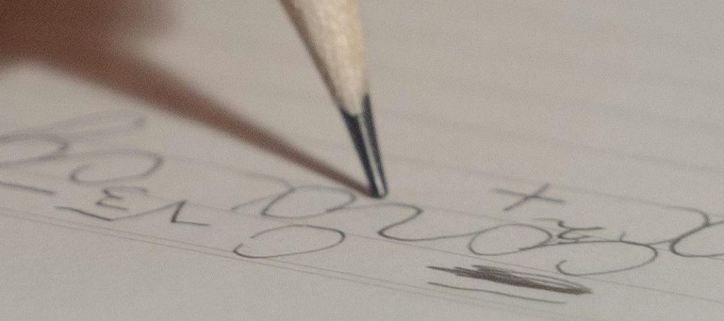 Um lápis escreve números e símbolos no papel, com traços irregulares e pouco consistentes, evidenciando dificuldade no controlo do movimento e na organização gráfica. A escrita apresenta variações no tamanho, alinhamento instável e pressão desigual, aspetos frequentemente associados à disgrafia. Este tipo de dificuldade pode refletir desafios na coordenação motora fina, no planeamento motor e na organização visuoespacial, interferindo na legibilidade e na fluidez da escrita.